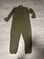 Front view of olive green flight jumpsuit laid flat showing full length, USA flag patch on arm, zippered pockets, and cuffed sleeves and ankles