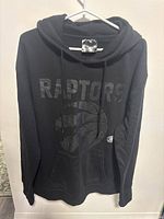 Front view of the black Toronto Raptors hoodie showing the logo and team name in gloss black lettering on matte black fabric.