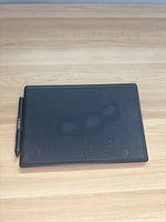 Top view showing black Wacom One drawing tablet with stylus pen on the left side, tablet surface shows light scratches and fingerprints.