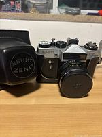 Front and side view of Zenit ET camera with lens attached and lens cap on, alongside protective case with Zenit logo on front.