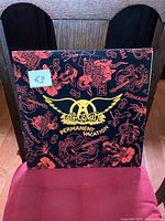 Front cover of Aerosmith Permanent Vacation LP album with black background and red illustrations plus yellow wings logo