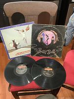 Two vintage vinyl records with their album jackets placed on a chair. Left jacket is stylized with a figure holding a guitar and a balloon dog, right jacket has hands holding a glowing orb with a dancing figure inside.