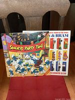 Photo showing the Smurfs Party Time album and the Sharon, Lois & Bram album with a poster placed behind them on a red surface, capturing the front covers.