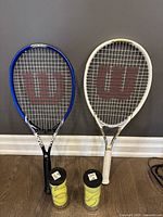 Two Wilson tennis racquets, blue Venus Serena and white Tova Slam, standing upright with two containers of tennis balls in front
