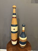 Two wooden pins or clubs, one larger with painted star and stripes, one smaller with blue and white painted stripes, shown together on wooden table