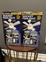 Photo of two boxed Bell + Howell Socket Fanlights side by side on a round wooden table. Box features product details and images showing the fanlight installed and functioning.