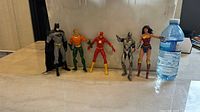 Five DC superhero action figures (Batman, Aquaman, The Flash, Cyborg, Wonder Woman) lined up on a counter with a water bottle for scale.