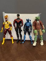 Four DC action figures with Nightwing, Beast Boy, Blue Beetle, and Firestorm lined up on a surface against a white background