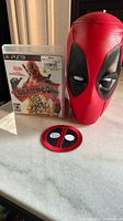Front view showing Deadpool PS3 game case, popcorn head, and logo patch on a table.