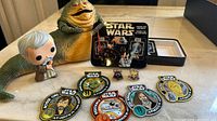 Photo showing large Jabba the Hutt figure, Funko Pop Obi-Wan figure, enamel pins, embroidered patches, and Star Wars playing cards in tin box.