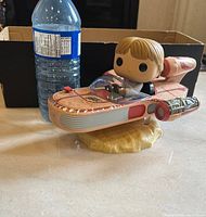 Front side view of Luke Skywalker Funko Pop figure seated in speeder with water bottle for scale and cardboard box in background.