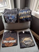 Four black Harley-Davidson t-shirts folded on chair showing front graphics and tags, illustrating the content and condition of the lot.
