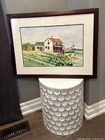 Framed watercolour painting showing a farmhouse and natural scenery, matted and set in a dark wood frame, placed on a white decorative stool against a wall.