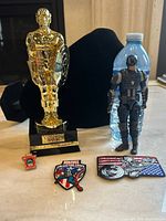 Five Marvel Captain America collectibles including gold statue on black base, Captain America action figure, and three embroidered pins on a table.