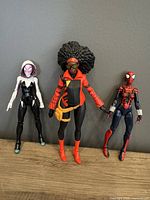 Front view of 3 Marvel Legends Spider-Women figures standing side by side on wooden surface against grey background.