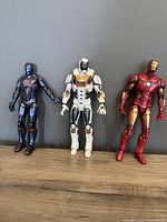 Front view of three Iron Man Marvel Legends figures in different suits against a gray wall and wooden surface.