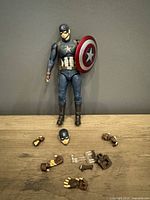 Full front view of Captain America figure with accessories laid out including shield, hands, additional head, and display stand parts.