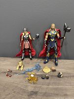 Front view of two action figures Thor and Jane Foster Thor standing with multiple accessories and interchangeable parts arranged below.