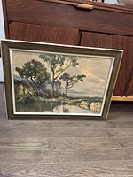 Framed signed print titled 'Still Waters' showing natural landscape with trees and reflection on water with muted colors.