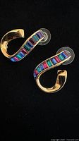 Pair of gold-tone wavy hoop earrings with rainbow baguette crystals and plastic backs on a black surface.