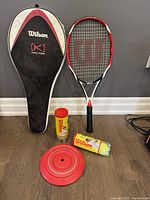 Wilson tennis racket, black and red with Wilson (K) Factor case, two packages of Wilson Championship tennis balls, and a red Tail Finz frisbee all on a wooden floor against a gray wall.