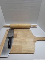 Set of three pizza-related kitchen tools: wooden pizza peel, pizza cutter with black handle, wooden rolling pin.