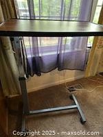 Full view of adjustable overbed table with woodgrain laminate top and metal base with casters next to window with curtains.