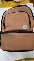 Front view of brown Carhartt backpack showing the brand patch on the front pocket with zipper closure.