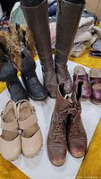 Photo showing nine pairs of assorted shoes including tall brown leather boots, black winter boots with fur lining, brown lace-up ankle boots, beige Mary Jane-style shoes, and dark purple clogs. Shoes placed on white surface for display.
