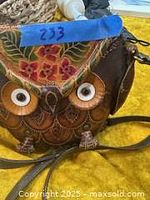 Close-up of the owl-themed brown handcrafted leather bag showing detailed feather tooling, large button eyes, and colorful floral accents on the flap.