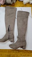 Pair of taupe over-the-knee boots made from suede with block heels featuring chain detail. The boots include a side zipper and pointed toe.