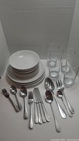 Complete modern dinnerware set for four with white plates and bowls, clear glasses, and stainless steel flatware displayed on table.