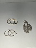Three pairs of silver earrings arranged on a flat surface: two pairs of medium-sized smooth polished hoops and one pair of double-leaf design dangle earrings