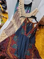 Three women's boho dresses hanging on hangers. Front view shows distinct colors and designs: navy and burgundy tribal print halter dress, tan dress with wide cream crochet straps and fringe, and green embroidered floral dress.
