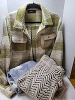Front view of the Blue B olive green and cream plaid sherpa shacket, gray flannel shirt partially folded, and beige chevron knit cardigan rolled up in front.