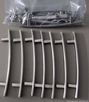 Overhead photo showing 13 curved brushed nickel handles laid out horizontally with mounting screws sealed in plastic bag above them