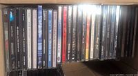 Row of assorted music CDs in jewel cases with various artist and album titles visible, arranged horizontally in a box.