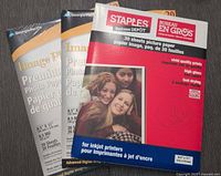 Two unopened packages of high gloss photo paper sheets for inkjet printers. One Staples Business DEPOT package with 30 sheets, red and blue packaging. One Image Plus Premium Photo Paper package with 20 sheets.