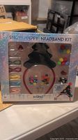 Front view of the unopened holiday DIY headband kit packaging showing a green felt Christmas tree headband base and assorted decoration pieces including beads and pom-poms