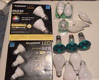 Photo showing 3 Luminus LED GU10 bulbs on original packaging and 2 Luminus PAR20 bulbs in original box, plus 3 green outdoor flood light bulbs and several white bulbs of chandelier and general use.