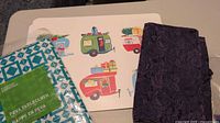 Six large plastic placemats with colorful camper van themed design and one purple damask table runner, displayed on a surface.