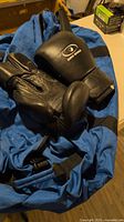 Budo World black sparring gloves resting on blue canvas gym bag, showing padded knuckles and wrist straps.