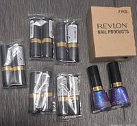 10 black and gold Revlon lipsticks in original sealed packaging, arranged in sets of two and three with one loose, next to a cardboard box containing 2 nail products.