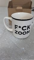 White ceramic mug with black rim, text 'F*CK ZOOM' on side, placed on bubble wrap with an open plain brown cardboard box behind it.
