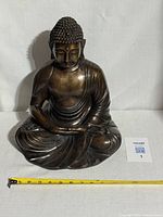 Front view of antique bronze Buddha statue seated in meditative posture, showing detailed robe and facial features. A measuring tape in front shows length for scale.