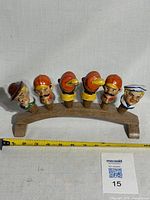 Full front view of the curved wooden stand with all six decorative porcelain wine bottle stoppers mounted and displayed, showing size reference with measuring tape.