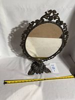 Antique brass vanity mirror with round reflective surface and ornate decorative frame and base, showing full front view with measuring tape for scale.