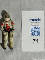 Front view of vintage 1983 GI Joe Ace V1 Fighter Pilot action figure in white space suit with red tubing and black gloves and boots, standing on white cloth background next to numbered card.