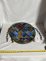 Front view of round stained glass panel showing mosaic colors and attached hanging chain laid on fabric background with measuring tape.