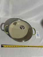 Top-down view of Le Creuset casserole pot showing glass lid with branded knob and light green handles, with measuring tape indicating approximately 11 inches diameter.